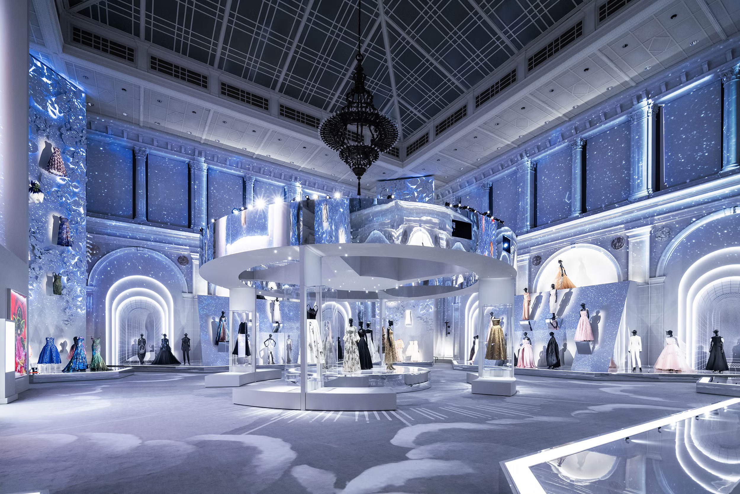 Luxurious fashion exhibit in an ornate hall with high ceilings, showcasing elegant gowns under soft blue lighting. A grand chandelier adds elegance.