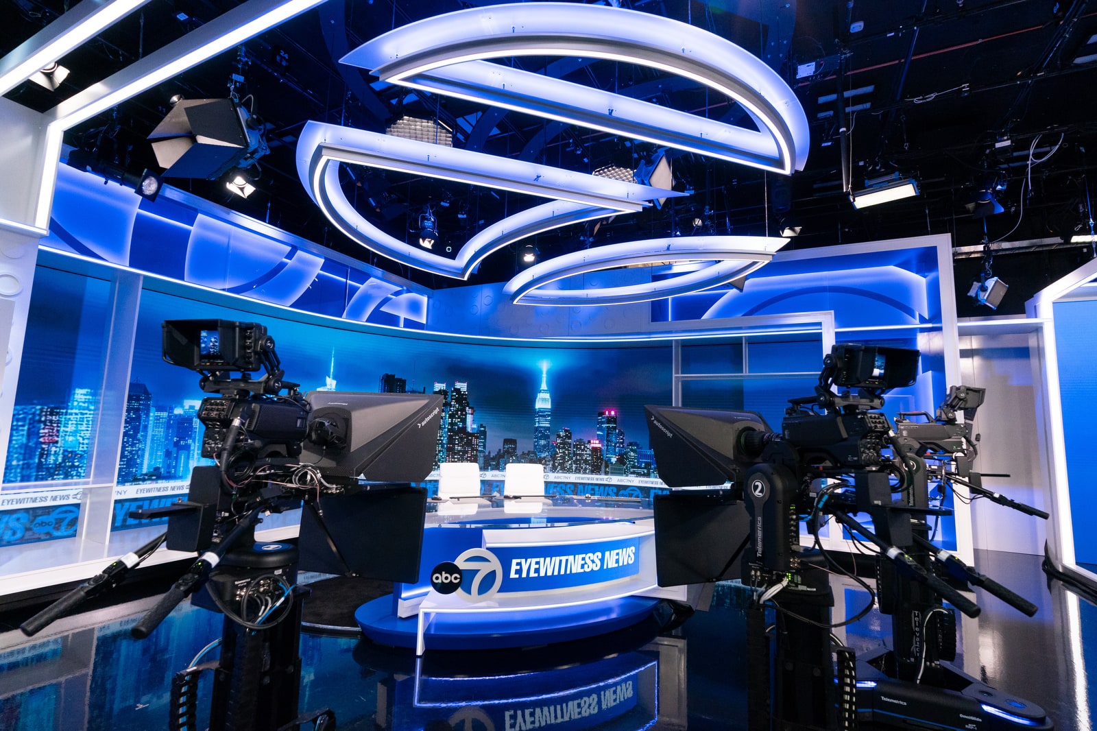 Modern TV news studio with blue lighting, large cameras, and two empty white chairs at a desk. A city skyline backdrop adds a dynamic atmosphere.